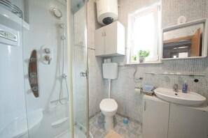 A bathroom with a glass-enclosed shower, a white toilet, a sink with a mirror above it, and a window with a view of greenery. - Apartments Kalajžić ST (Pucisca)