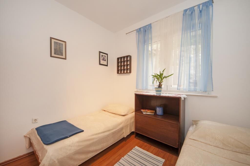 A small, tidy room with two single beds, a wooden nightstand, a window with sheer curtains, and a small shelf with a plant.