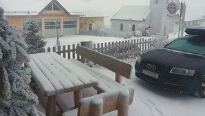 A snow-covered outdoor seating area with a bench and a table, a car parked nearby, and a building with a sign reading "FEUERWEHR BRAUNEGG" in the background. - Brauneggerhof Gasthof Mayer (Raxendorf)