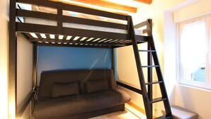 Una cama litera con escalera, un sofá y una ventana en una habitación con vigas de madera y suelo de color claro.