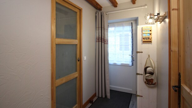 Un pasillo estrecho con vigas de madera, una puerta de vidrio esmerilado, una ventana con cortinas y un zapatero con pantuflas.