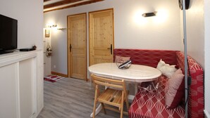 Una sala de estar acogedora con un sofá rojo, una mesita redonda pequeña y un televisor sobre una repisa blanca. Se observan vigas de madera y una lámpara en el techo.