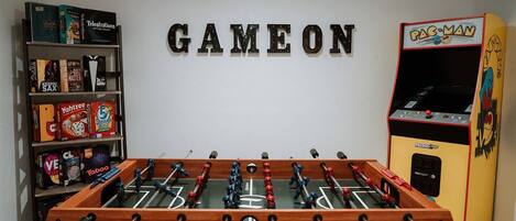 A room with a foosball table, a Pac-Man arcade machine, and a bookshelf with various board games.