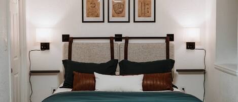 A bedroom with a bed, headboard, bedside lamps, and three framed pictures on the wall.