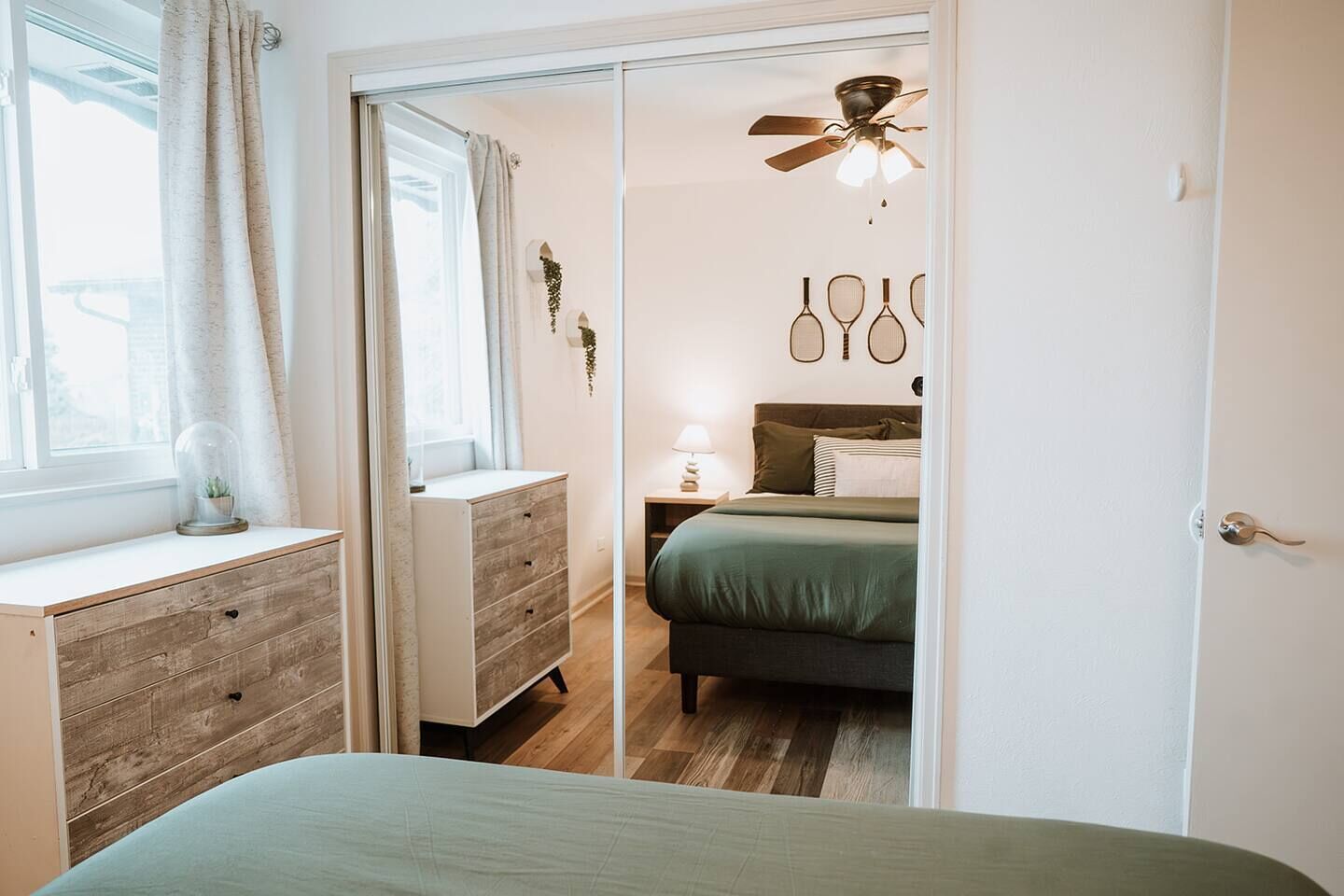 Un dormitorio con cama, mesita de noche, cómoda, ventana con cortinas y ventilador de techo.