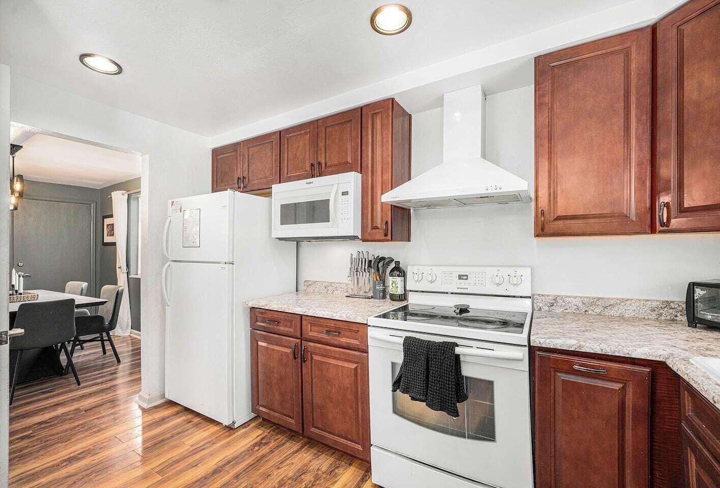 A modern kitchen with wooden cabinets, a white refrigerator, a microwave, a stove, and a dining area with a table and chairs.