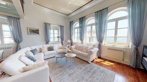 Un salon spacieux avec un grand canapé en forme de L, une table basse et un téléviseur. De grandes fenêtres sont agrémentées de rideaux, un radiateur est installé et un tableau orne le mur.