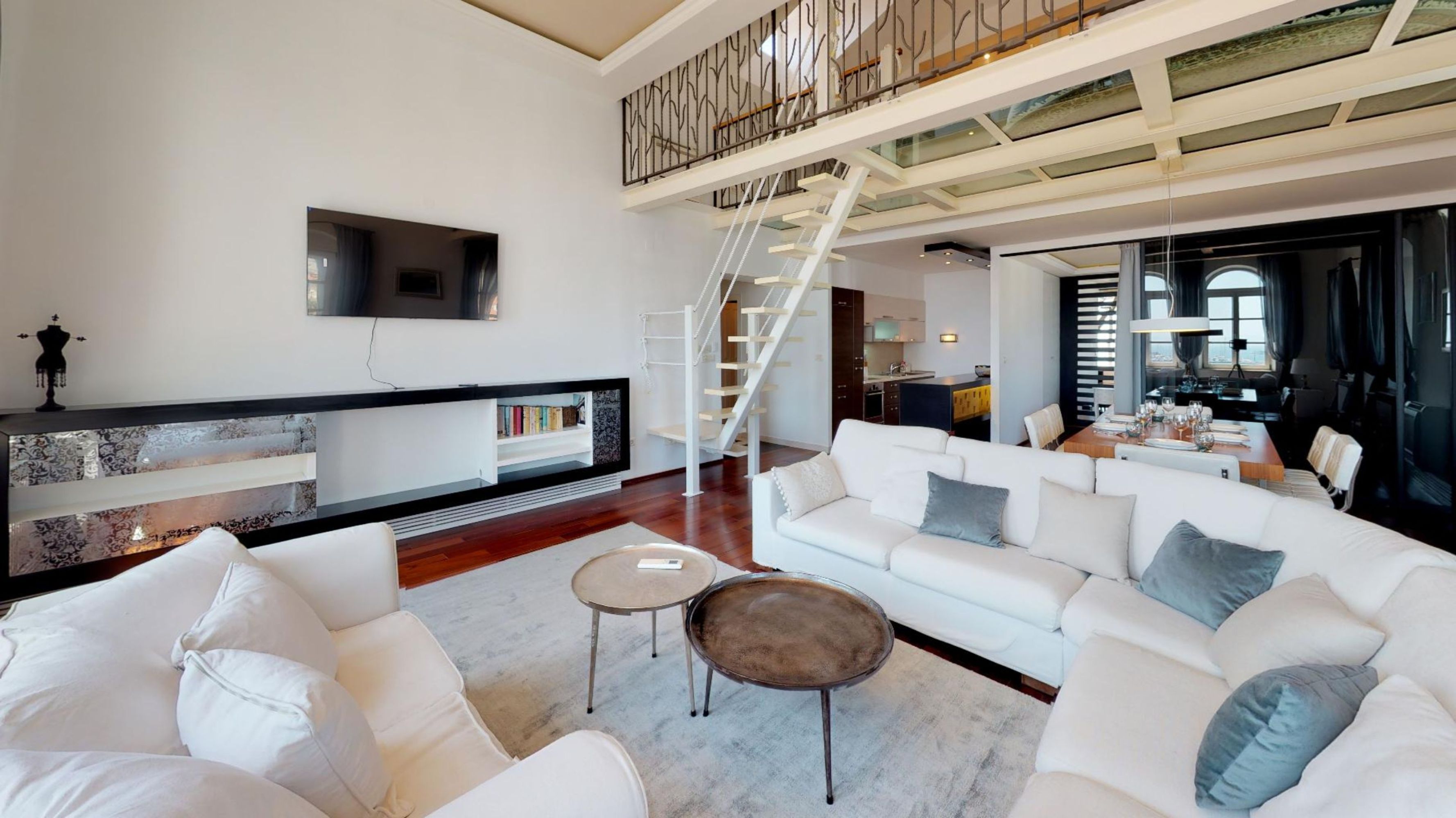 A spacious living area with a white sofa set, a coffee table, and a TV mounted on the wall. A staircase leads to an upper level with a balcony.
