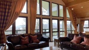 A spacious living room with large windows, leather sofas, a wooden coffee table, and a lamp. - Luxury Loch Lomond Lodge (Alexandria)