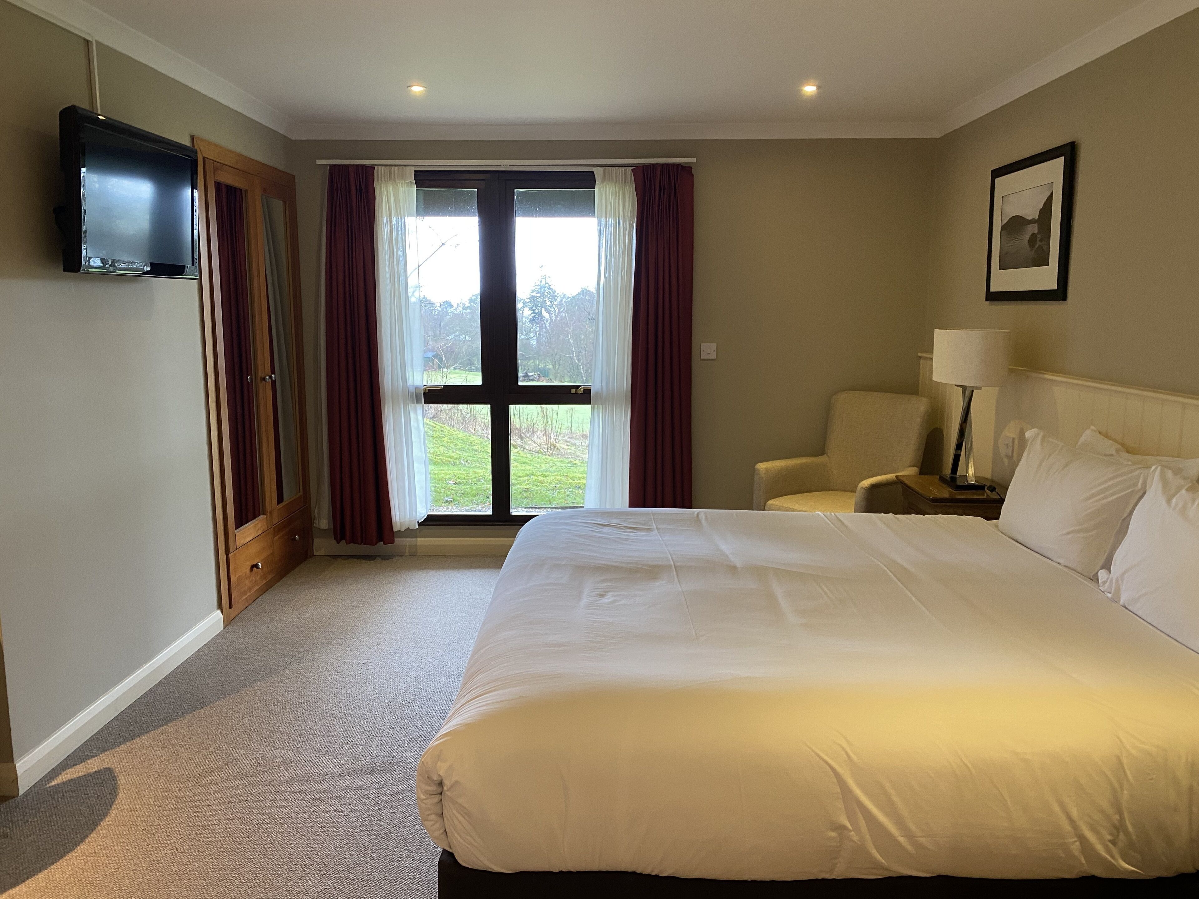 Une chambre d’hôtel avec un grand lit, une chaise, un bureau avec une lampe, une télévision et une fenêtre avec des rideaux.