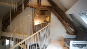 Een slaapkamer in een loft met een schuine kap, zichtbare houten balken, een spiltrap en een bed met hoofdeinde.