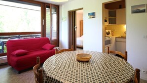 Une pièce chaleureuse avec un canapé rouge, une table ronde recouverte d’une nappe à motifs et un coin cuisine visible à travers une porte ouverte.