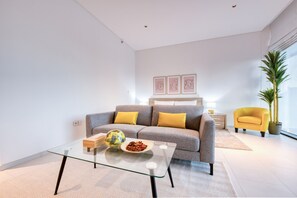 A modern living room with a glass coffee table, a grey sofa with yellow cushions, a yellow armchair, and a large potted plant. - Stylish Studio w/ Dubai Canal Vw in Business Bay (Dubai)