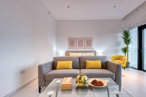 Un salon moderne avec un canapé gris, des coussins jaunes, une table basse en verre et des œuvres d’art encadrées accrochées au mur.