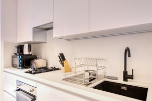 A modern kitchen with white cabinetry, a stainless steel sink, a black faucet, a toaster, a coffee maker, and a knife holder. - Stylish Studio w/ Dubai Canal Vw in Business Bay (Dubai)