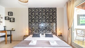 Una cama bien tendida con sábanas blancas y dos almohadas, una mesita de noche con una lámpara y un tapiz en la pared.