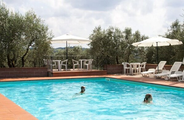 A swimming pool with two people in the water, surrounded by white lounge chairs and umbrellas.