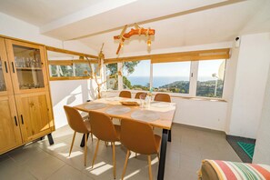A dining area with a table set for four, a wooden cabinet with glass doors, and a view of the sea through large windows. - Villa Vista | Amazing Sea Views Pool BBQ Air Conditioning Ideal for Families (Tossa de Mar)