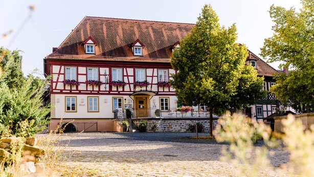 Un bâtiment traditionnel à colombages, doté d’une cour pavée, d’une porte en bois et de jardinières aux fenêtres.