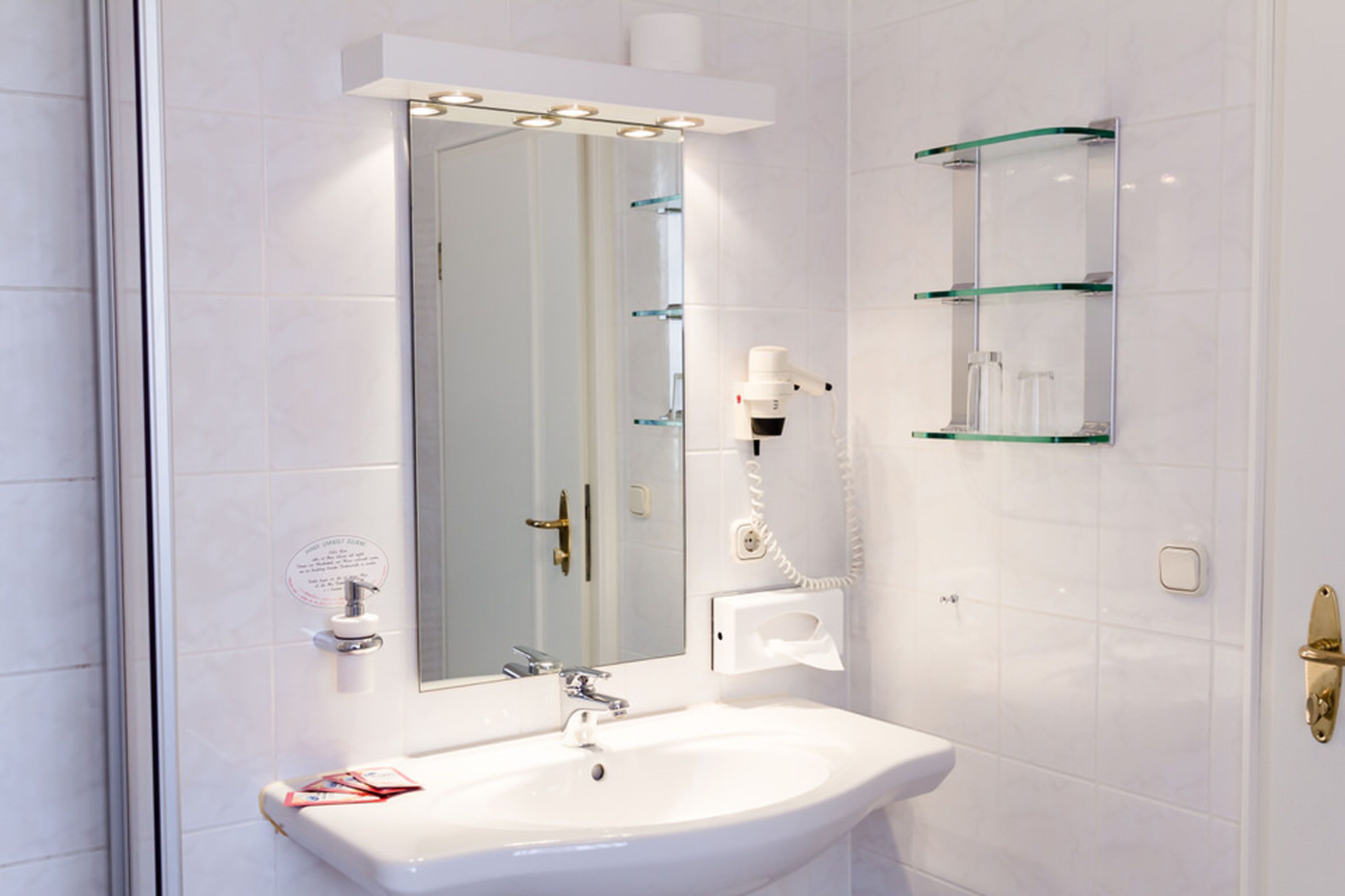 A bathroom with a white sink, a mirror, and a glass shelf unit.