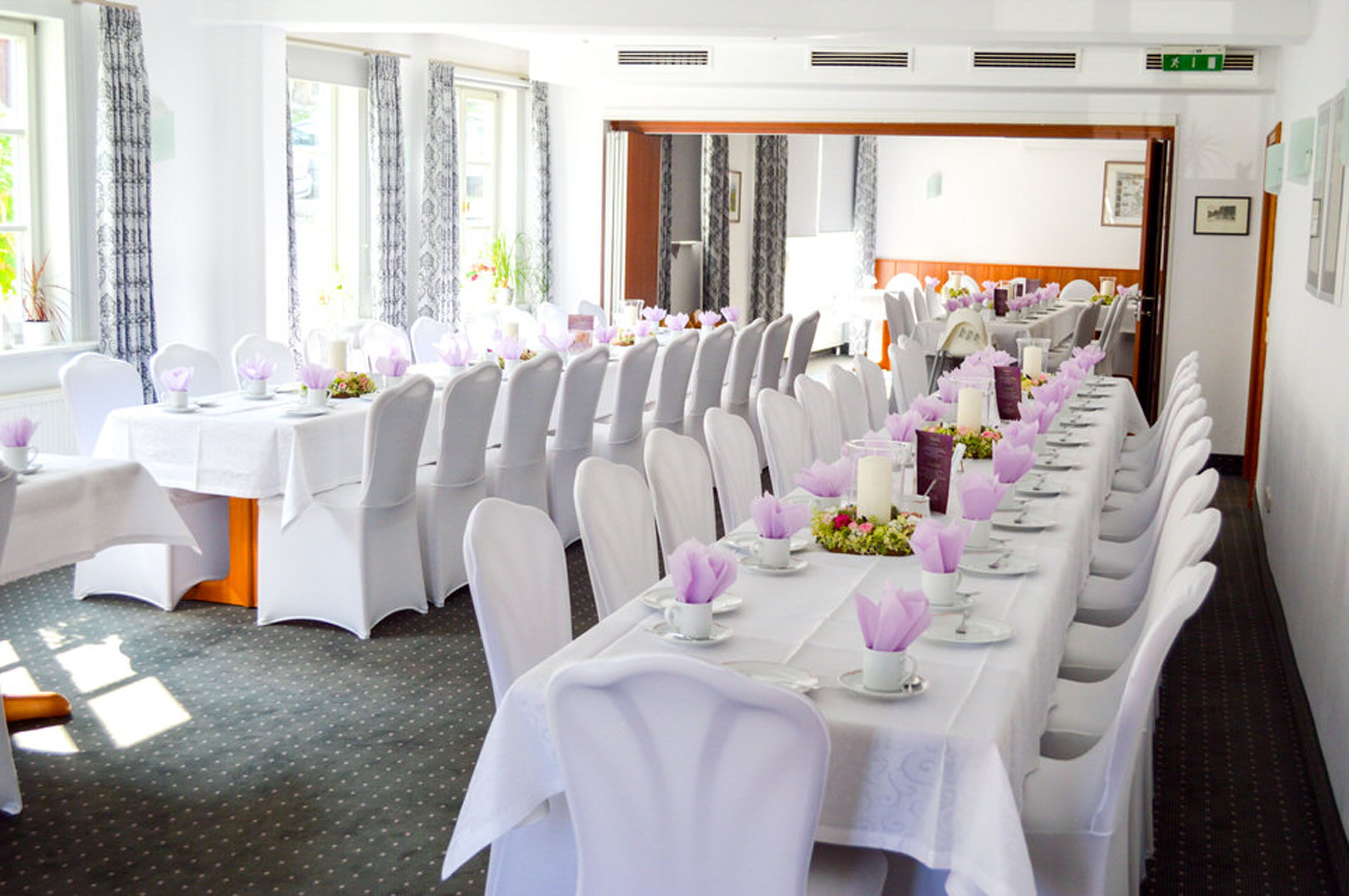 Ein Festsaal mit langen Tischen, festlich gedeckt für eine Veranstaltung, mit weißen Tischdecken, Stühlen und floralen Tischdekorationen.