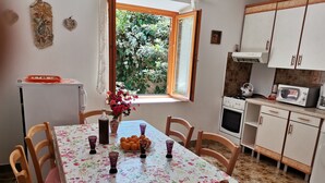 Ein Essbereich mit Tisch und vier Stühlen, umgeben von Holzsesseln. Ein Fenster mit Blick ins Grüne ist erkennbar, und eine angrenzende Küche mit Schränken und Geräten.