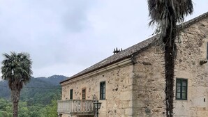 Un edificio de piedra con un balcón, rodeado de vegetación y montañas al fondo.