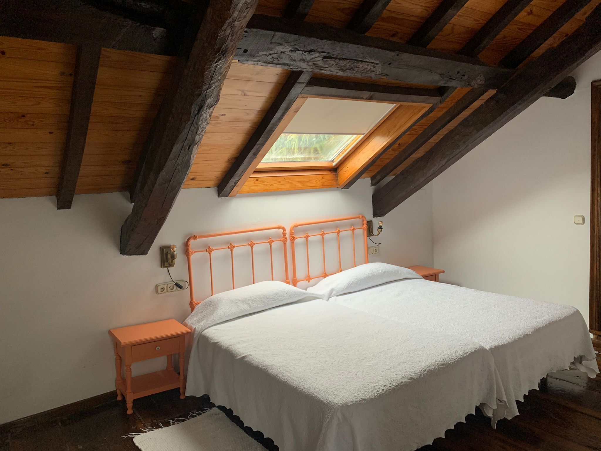 A bedroom with a sloped ceiling, a skylight, a wooden bed frame, and a small bedside table.