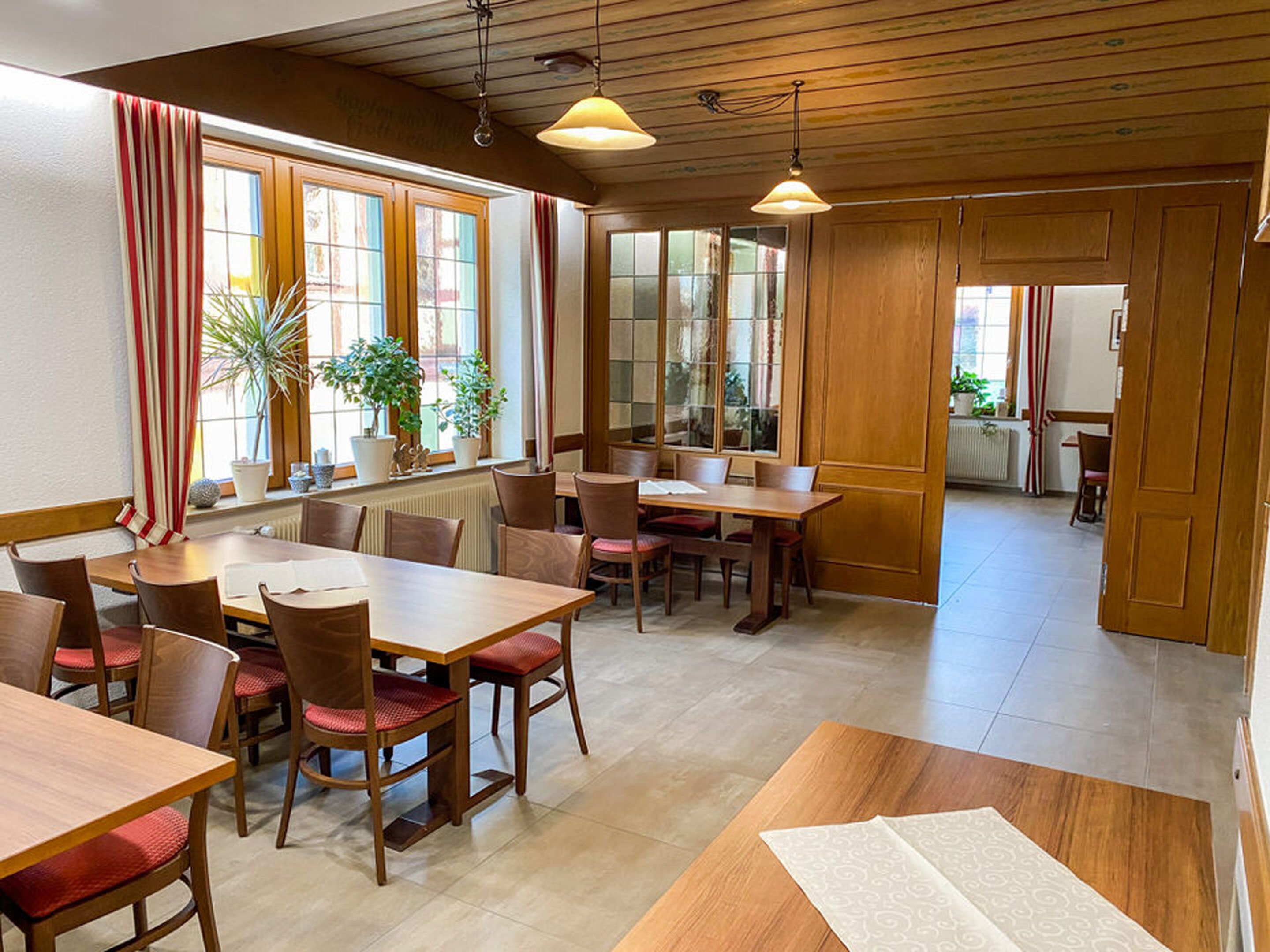A dining area with wooden tables and chairs, large windows with curtains, and potted plants.