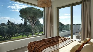 A bedroom with a large window, a bed with a brown blanket, and a view of trees and greenery.