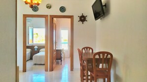 A room with a wooden dining table and chairs, a TV mounted on the wall, and a door leading to another room with a bed and a window. - Piso a pie de Playa (Suances)