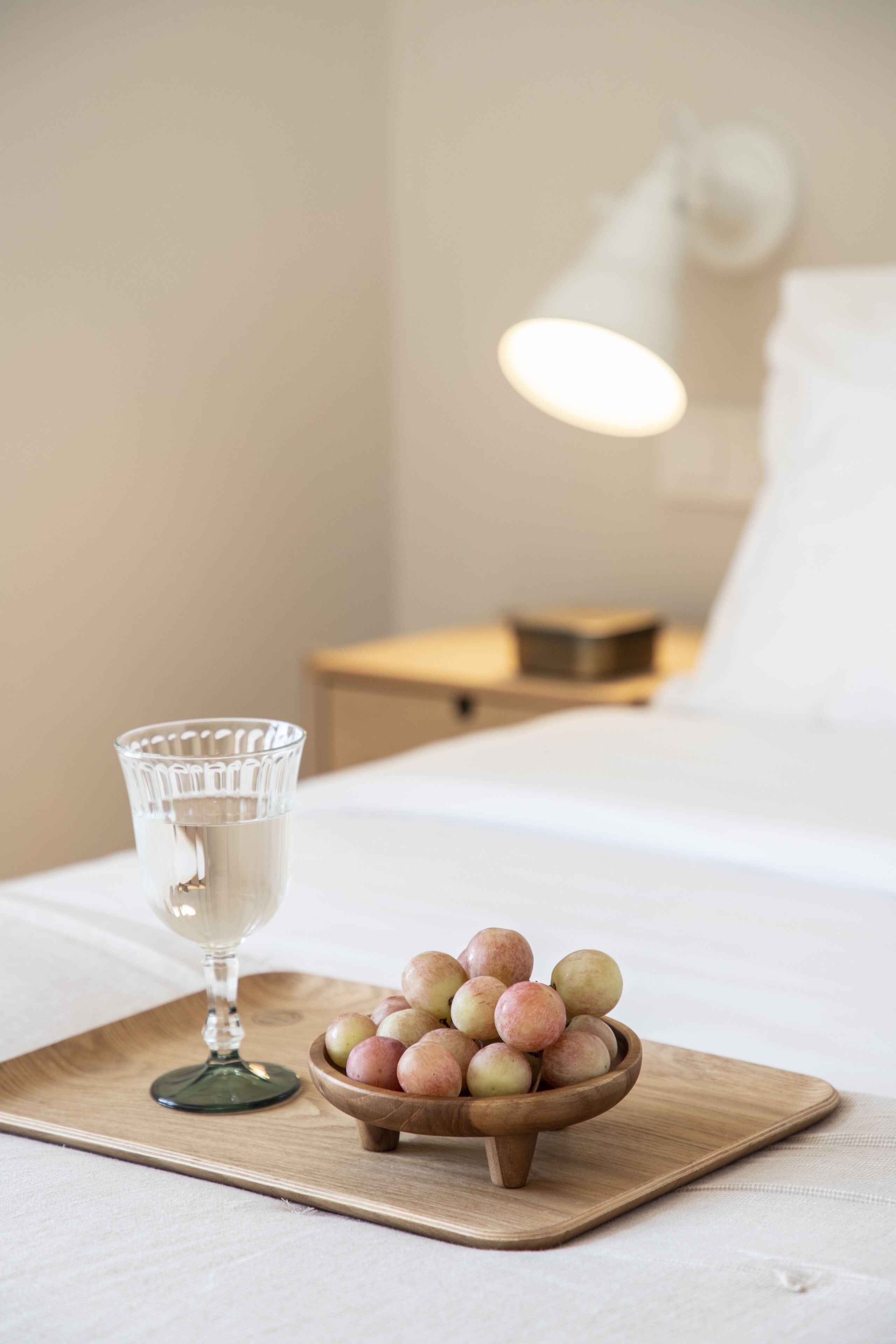 A wooden tray with a glass and a bowl of fruit on a bed.