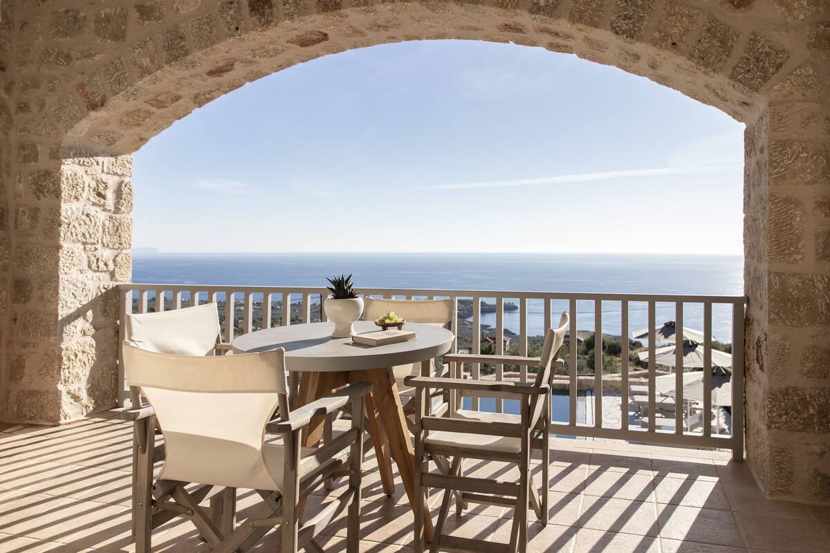 A terrace with a stone archway, a round table, and chairs, overlooking a body of water.