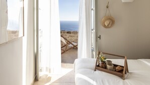 A bedroom with a view of the sea, a straw hat on the wall, and a tray with a vase and mug on the bed. - Amani Earthy Living (West Mani)