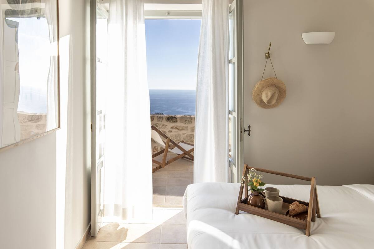 A bedroom with a view of the sea, a straw hat on the wall, and a tray with a vase and mug on the bed.