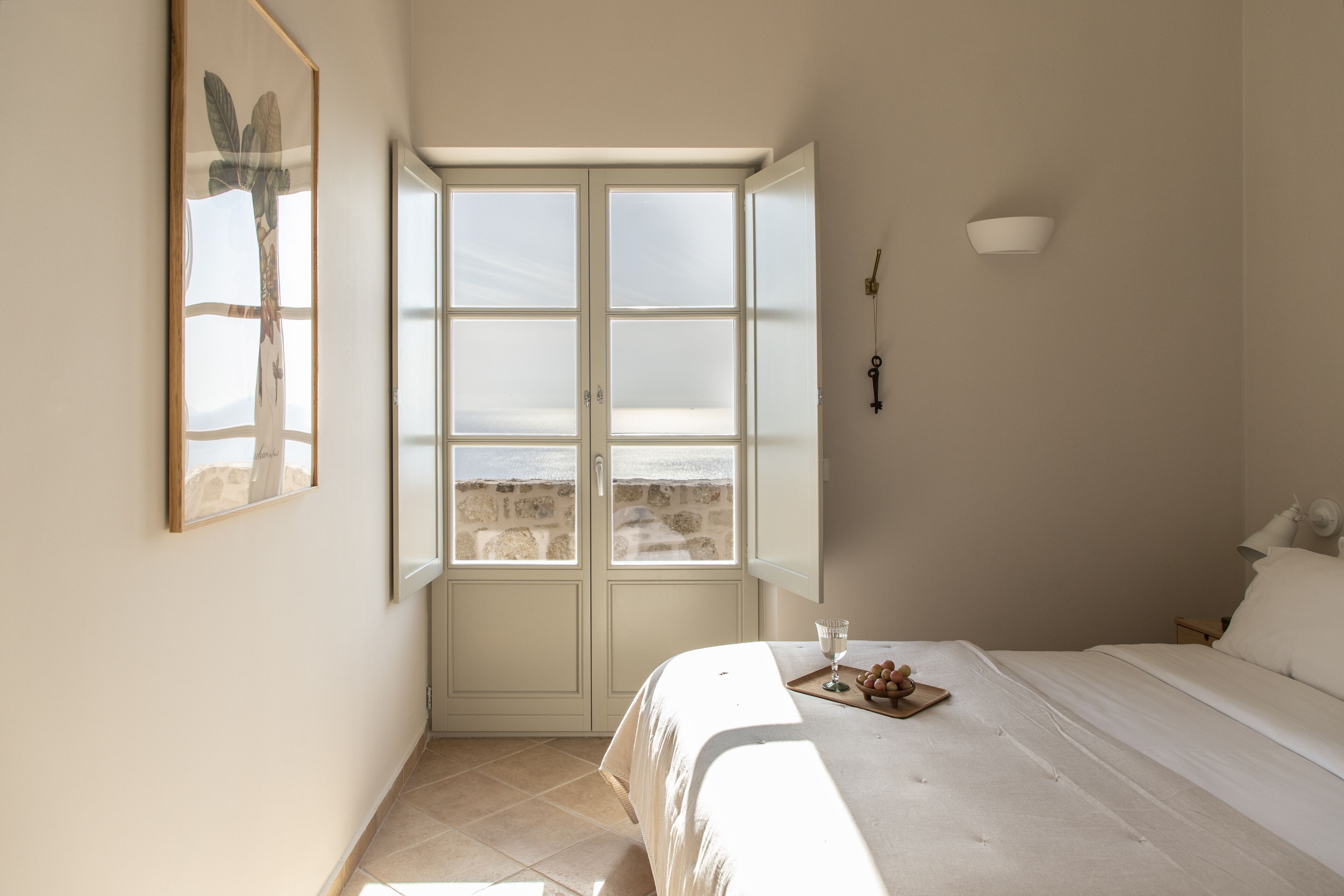 A bedroom with a large window, a bed, a bedside table with a glass, and a framed picture on the wall.