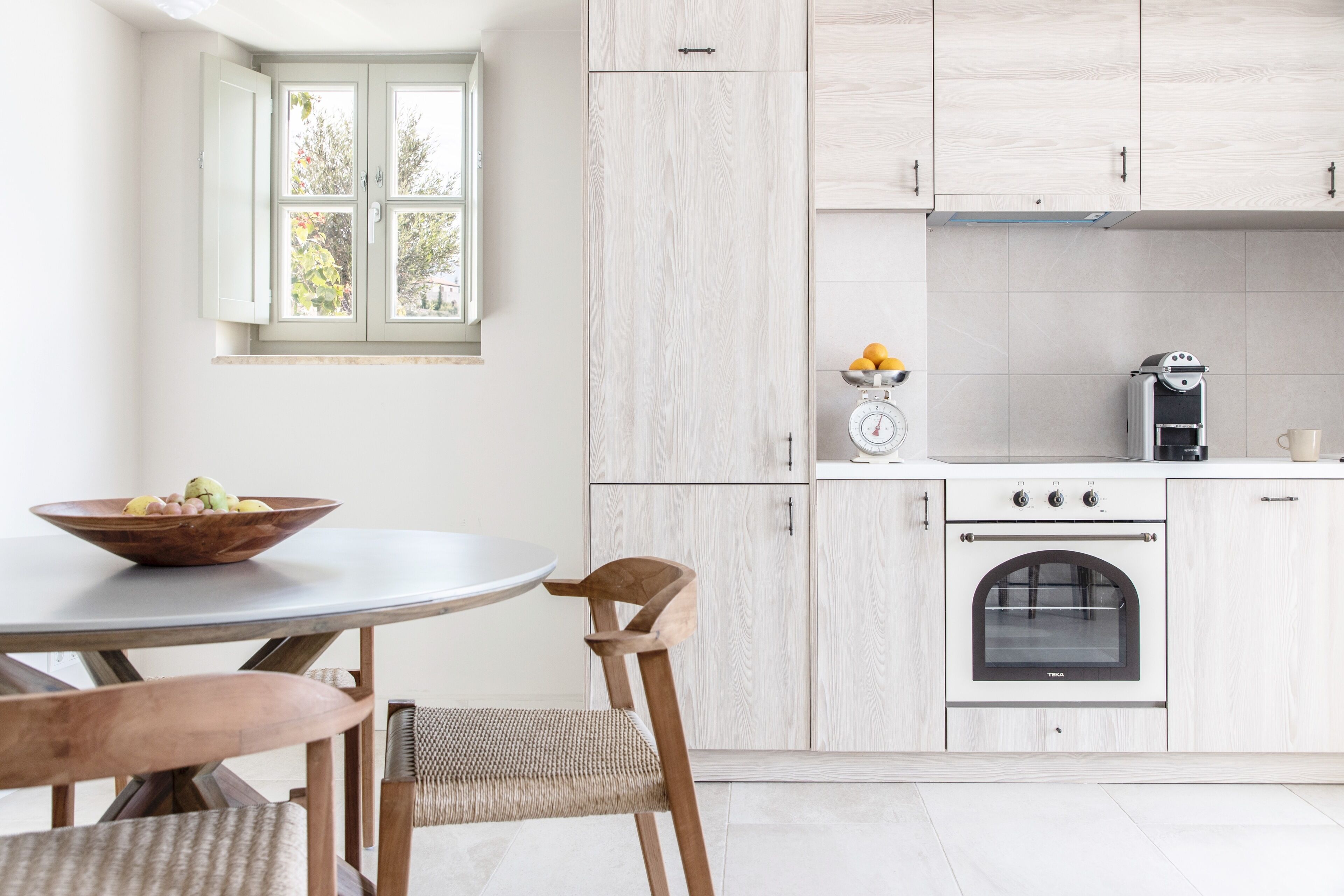 A modern kitchen with a round table, two chairs, a bowl of fruit, a window, and a stove.