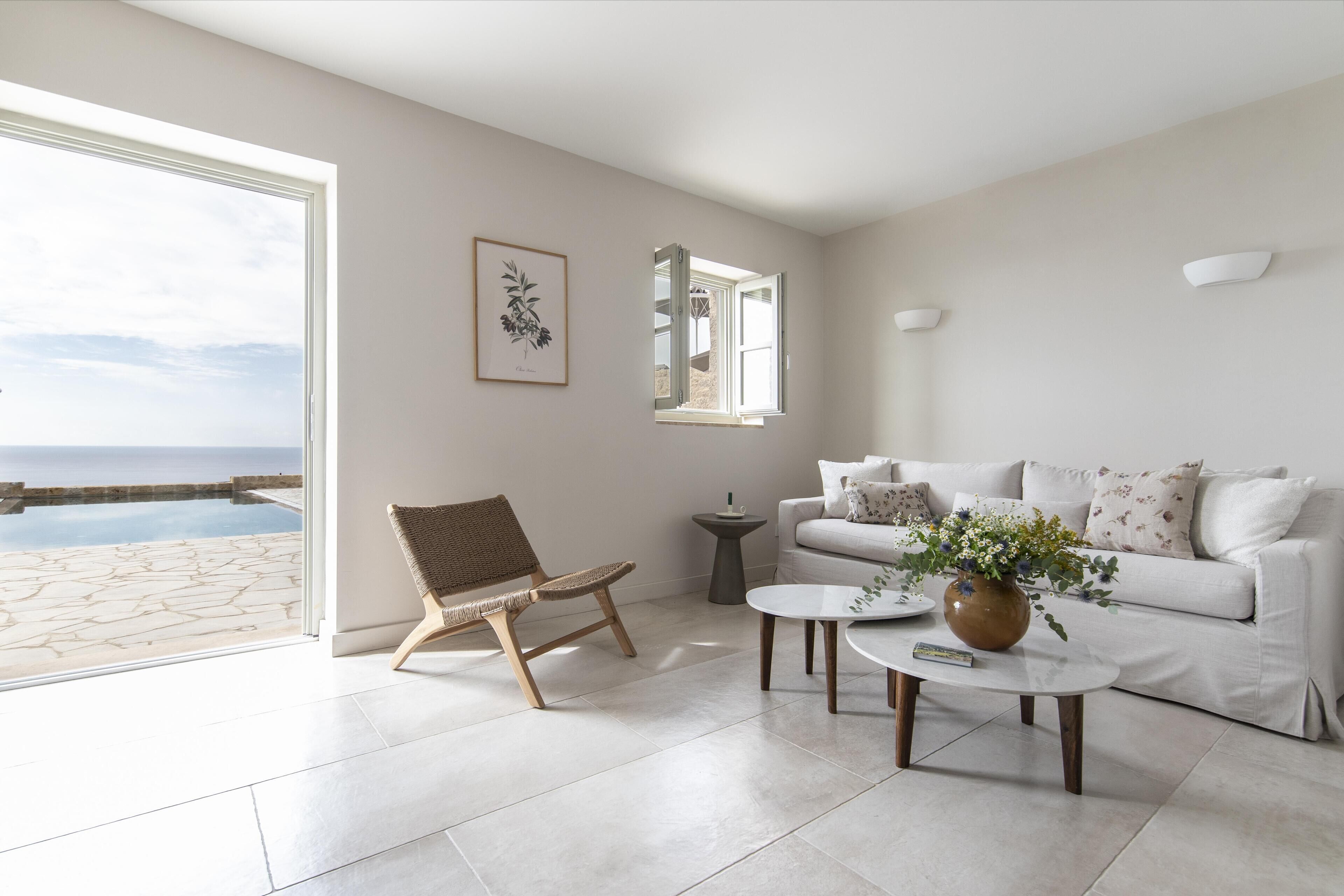 A modern living room with a white sofa, a wicker chair, a coffee table, and a view of the ocean through a large window.
