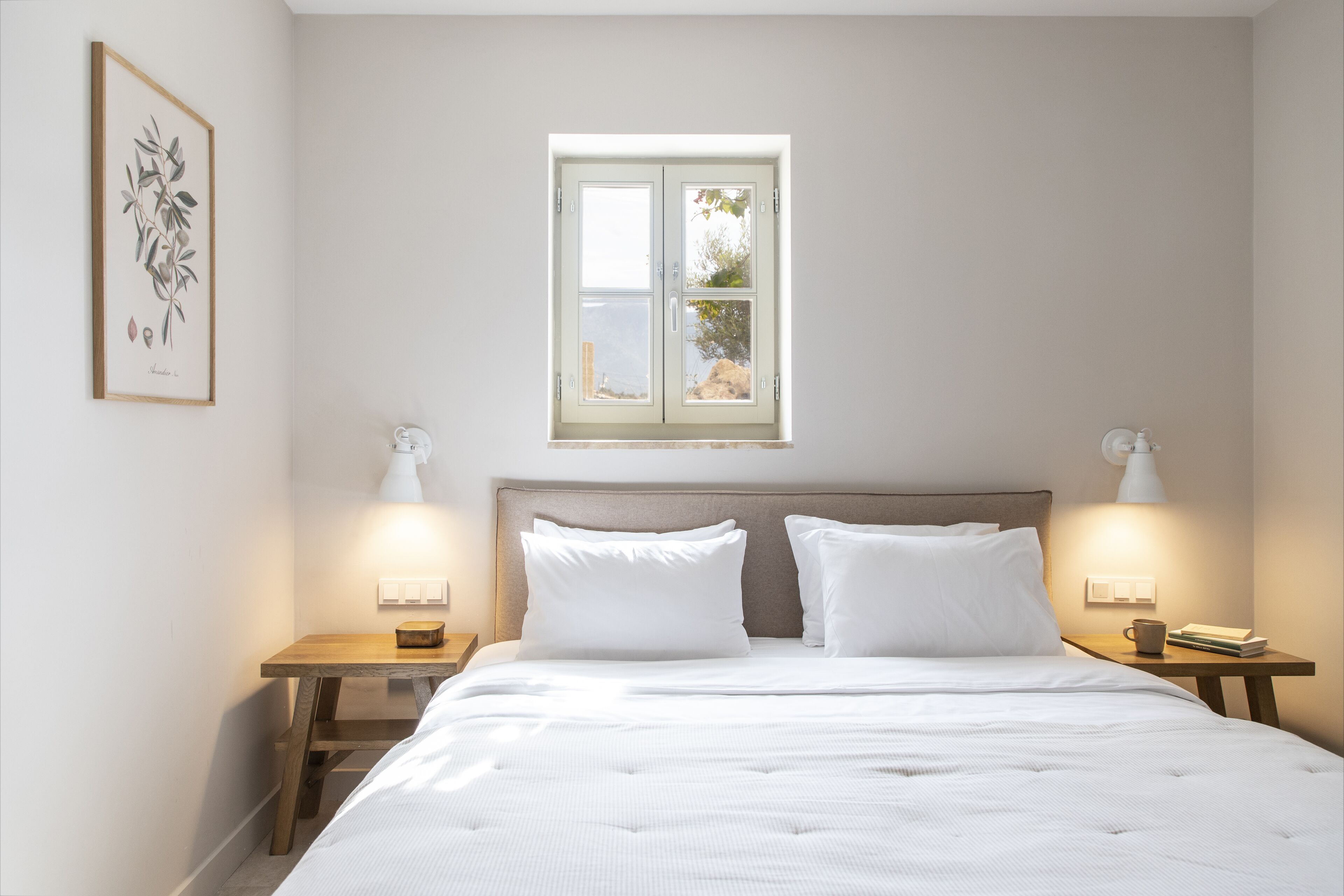 A neatly made bed with white linens, two bedside tables with lamps, a framed botanical print on the wall, and a window with a view of greenery.