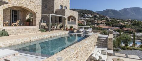 Un edificio de piedra con piscina, mobiliario exterior y vistas a un pueblo en la ladera.