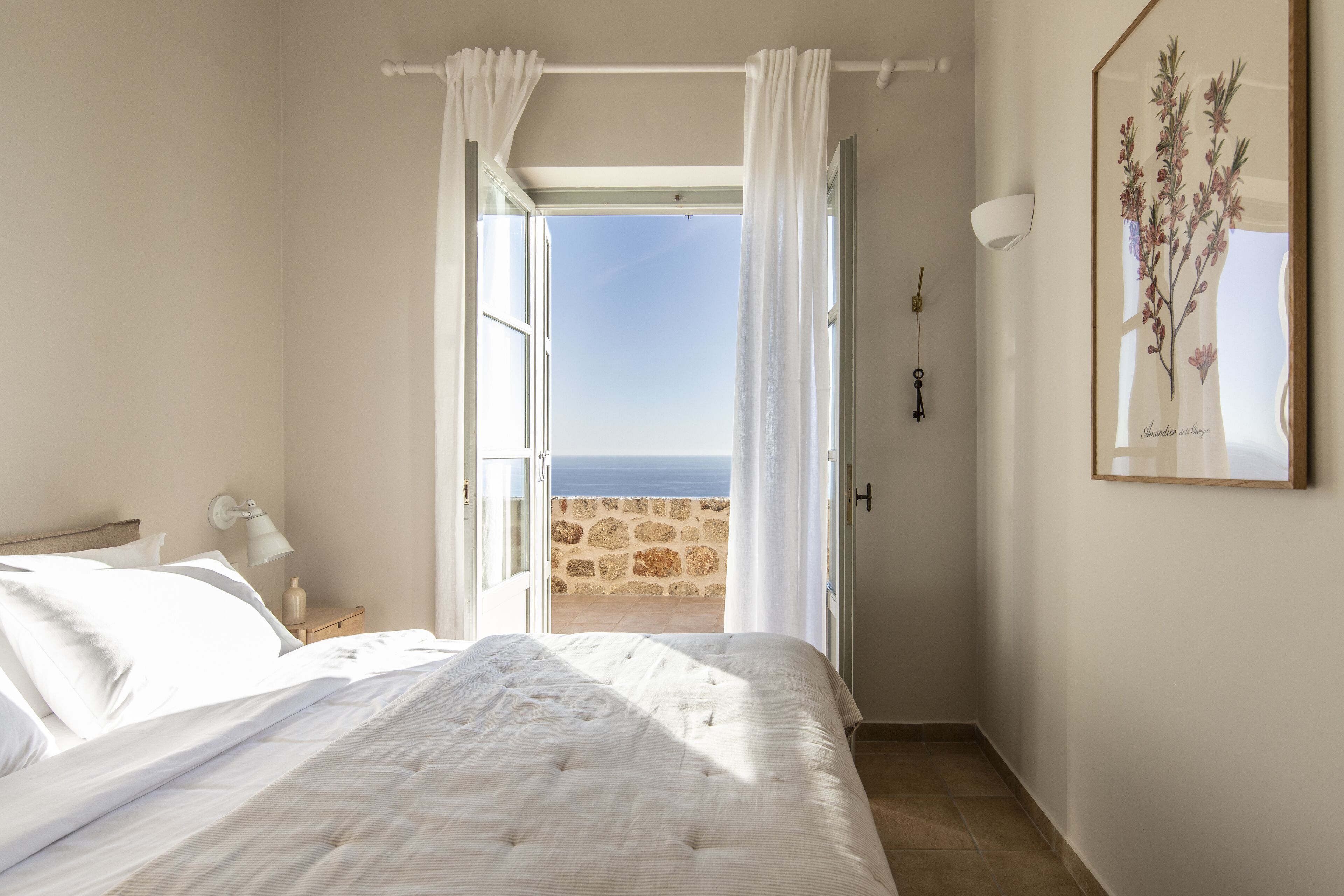 A bedroom with a bed, a nightstand, a lamp, a framed botanical print, and a view of the sea through an open door.