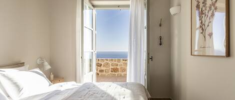 A bedroom with a bed, a nightstand, a lamp, a framed botanical print, and a view of the sea through an open door.