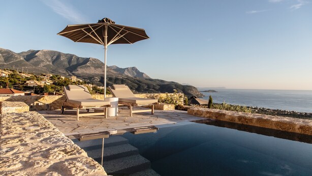 An infinity pool with lounge chairs and an umbrella, overlooking a coastal landscape with mountains in the background. - Amani Earthy Living (West Mani)