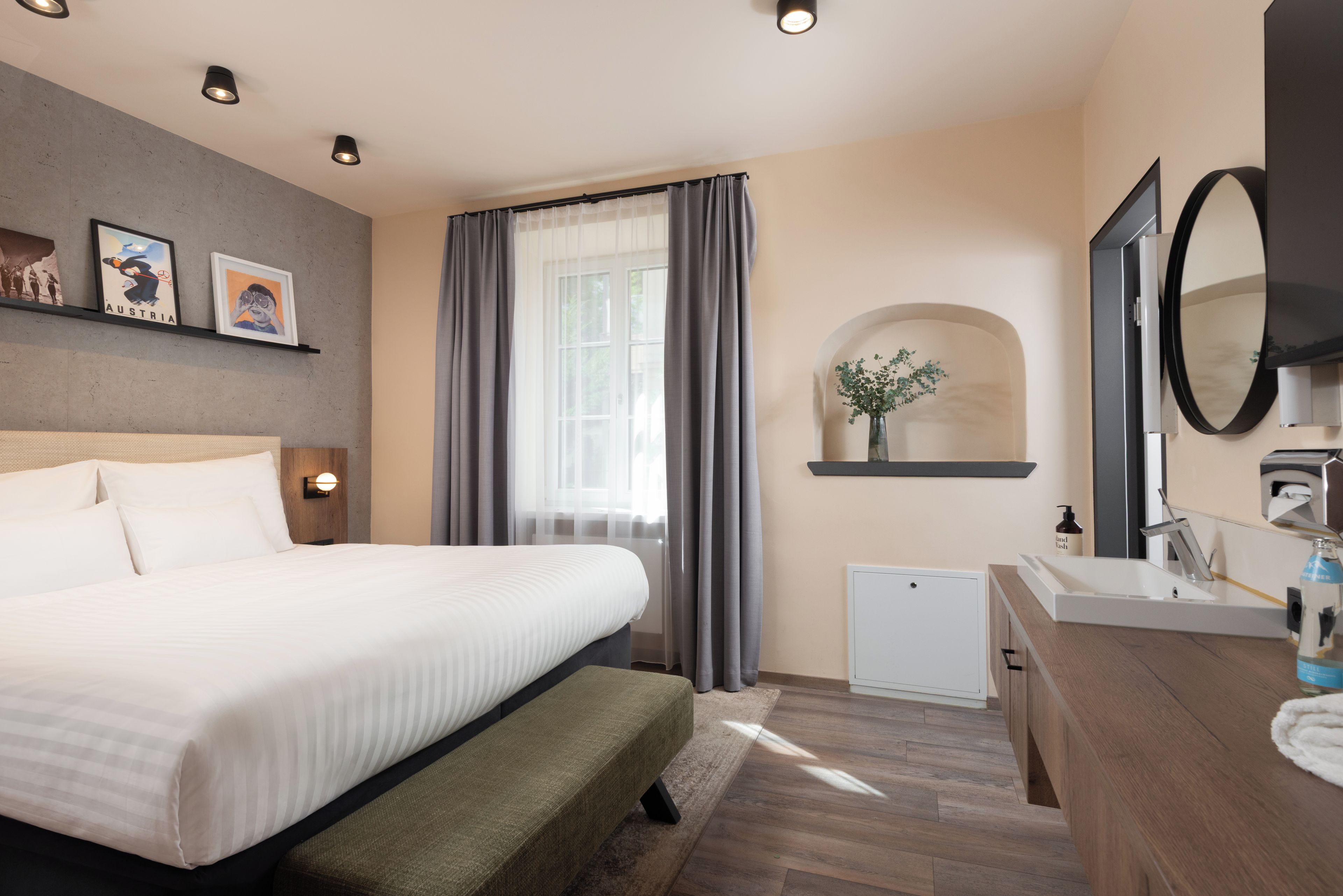 Habitación de hotel con cama, mesita de noche, un pequeño escritorio con silla, un espejo, ventana con cortinas y estante en la pared con una planta.