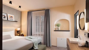 Habitación de hotel con cama, mesita de noche, lámpara, ventana con cortinas y baño con lavamanos y espejo.