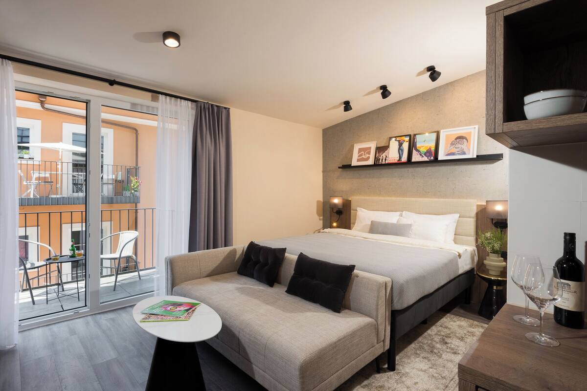 A modern hotel room with a bed, a sofa, a small table, and a balcony with chairs.