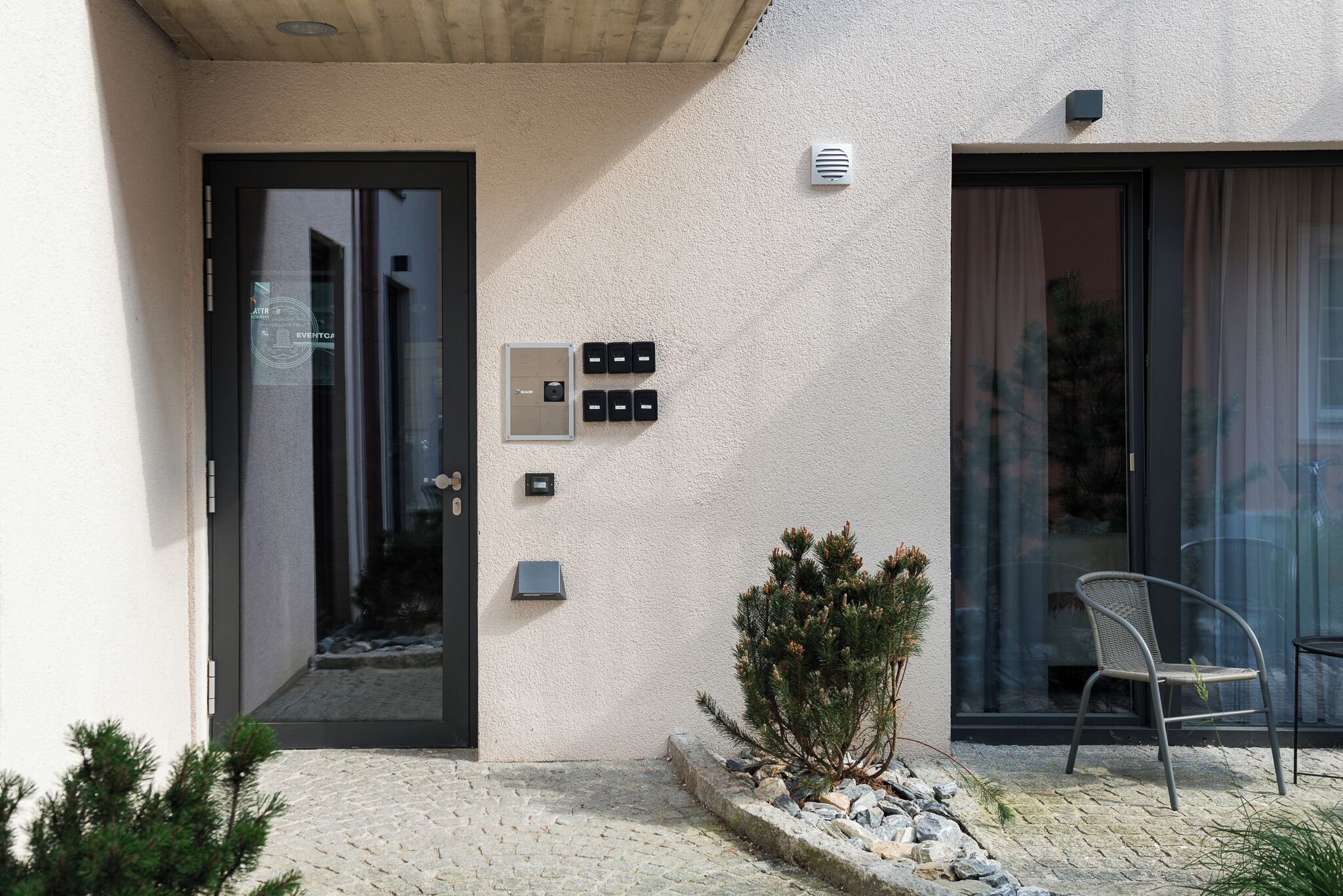 A building entrance with a glass door, a small potted plant, a chair, and a stone pathway.