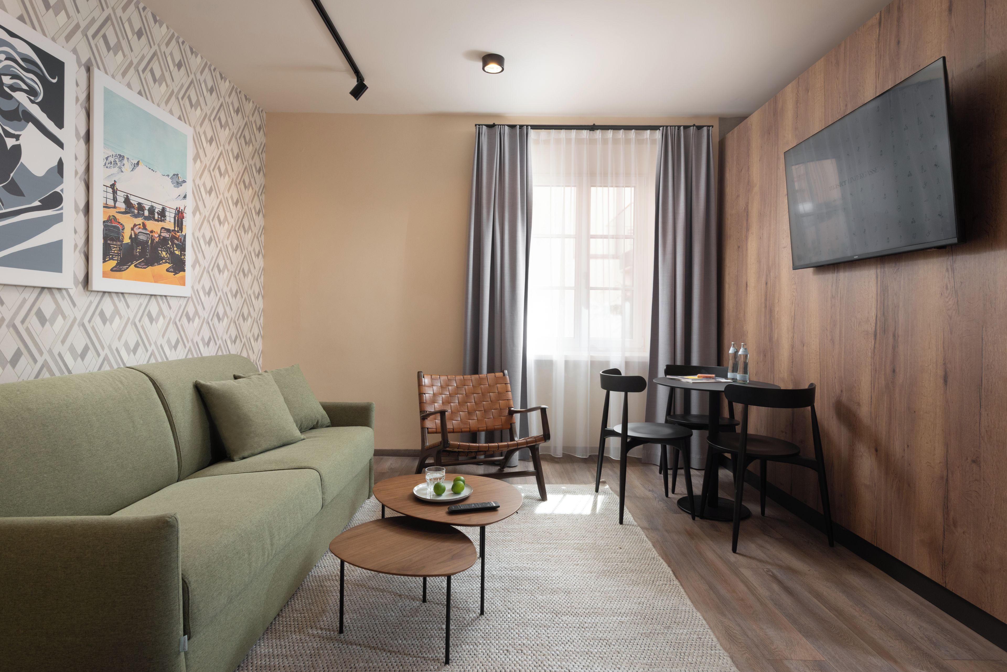 A modern living room with a green sofa, a coffee table, a dining table with chairs, a TV mounted on the wall, and a window with curtains.