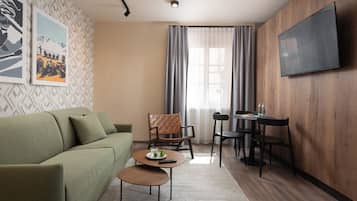 A modern living room with a green sofa, a coffee table, a dining table with chairs, a TV mounted on the wall, and a window with curtains.