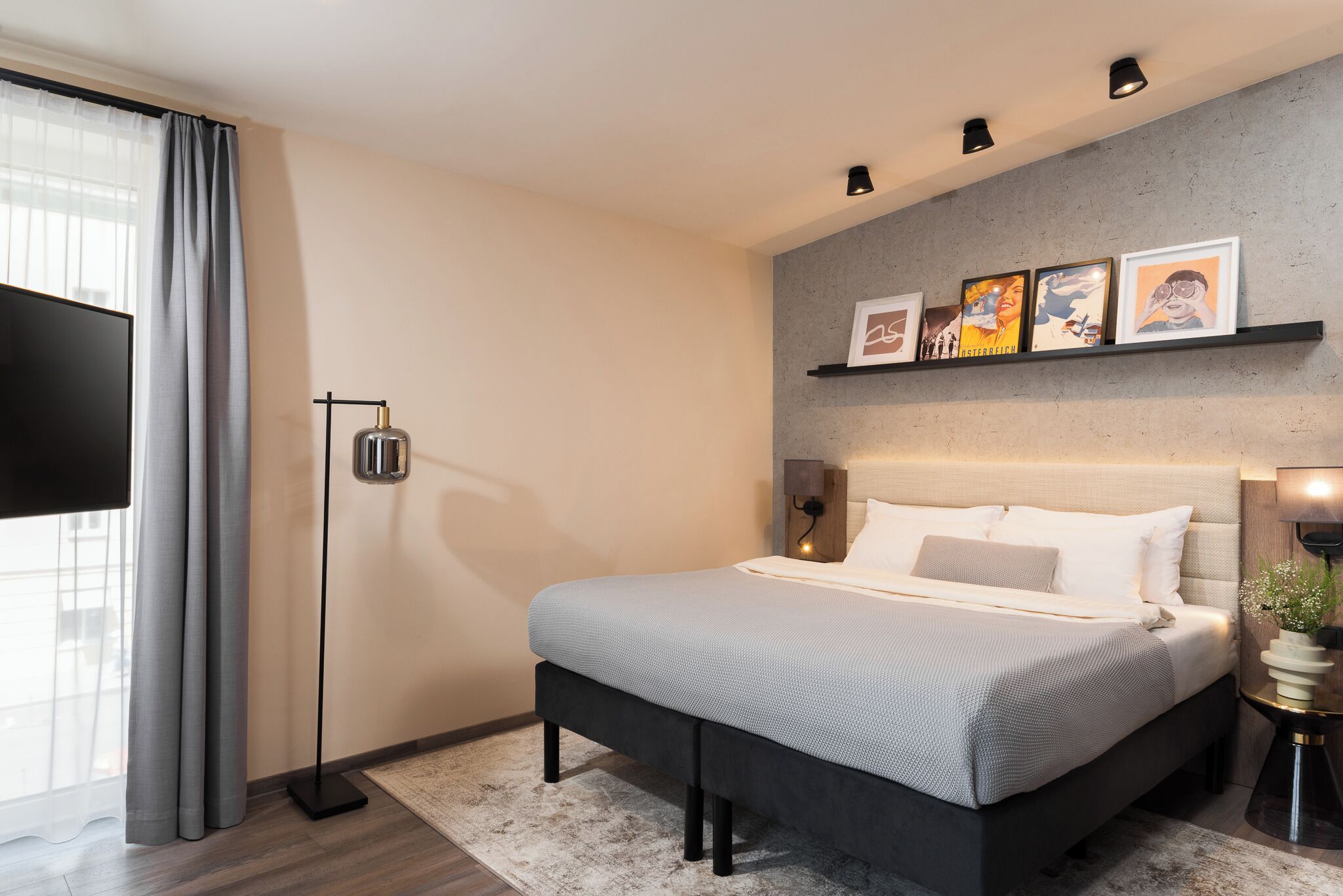 Un dormitorio moderno con una cama grande, un televisor de pantalla plana, una lámpara de pie y un estante de pared con obras de arte enmarcadas.